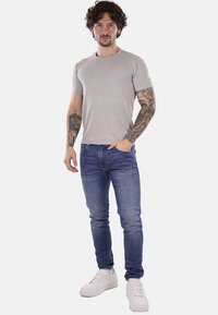 Camiseta corta de manga gris con una superficie texturizada, combinada con jeans ajustados azules y zapatillas blancas. Presenta tatuajes visibles en los brazos.