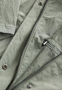 Light green, textured fabric jacket with a front zipper, snap buttons, and an interior pocket. Features a slightly crinkled appearance.