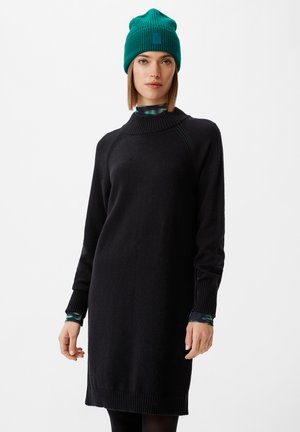 Schwarzes Strickkleid mit hohem Kragen und gerippten Bündchen, kombiniert mit einer grünen gerippten Mütze mit Logo-Patch. Model trägt schwarze Leggings.