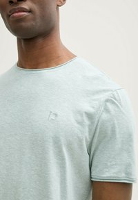 Camiseta de algodón verde claro con cuello redondo, mangas cortas y un sutil logo en relieve en el pecho. Tejido suave y texturizado.