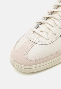 Zapatilla con cuero blanco y acentos de ante beige claro, que presenta una puntera redondeada, detalles cosidos y suela de goma texturizada.