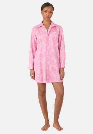 Vrouw staat op blote voeten en draagt een lange mouw, roze jurk met paisley-patroon, kraag en knopen aan de voorkant.