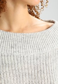 Maglione grigio chiaro lavorato a maglia con un ampio scollo a coste, caratterizzato da un motivo testurizzato e una vestibilità rilassata, visto dalla spalla in su.