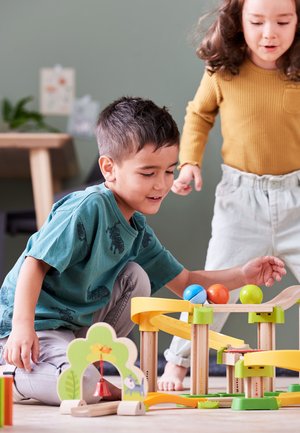 Buntes Holz-Kugelbahn-Spielzeug mit gelben und grünen Schienen, drei Kugeln (blau, orange, grün) und dekorativen Elementen auf einem hellen Holzboden.