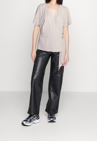 Top beige de manga corta con cierre tipo envoltura, combinado con pantalones de cuero negros y zapatillas azul marino. El top presenta un cierre con lazo y un diseño de escote en V.