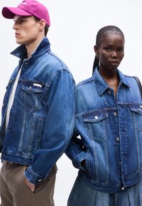 Άνδρας με ροζ καπέλο και γυναίκα με πλεγμένα μαλλιά, και οι δύο φορούν μπλε τζιν μπουφάν, στέκονται δίπλα-δίπλα σε λευκό φόντο.