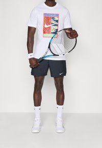 Camiseta blanca de Nike con diseño colorido de animales, pantalones cortos negros, calcetines blancos y zapatillas de tenis blancas, sosteniendo una raqueta de tenis.