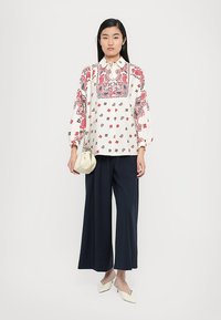 Vrouw in een witte en rode gebloemde blouse, met wijde zwarte broek, witte slingback hakken en een kleine witte handtas.