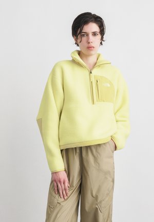 Persoană îmbrăcată într-un pulover ușor galben din fleece cu buzunar cu fermoar pe piept și pantaloni cargo bej, stând în fața unui fundal alb simplu.