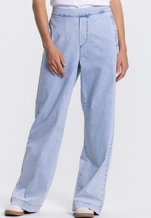 Vrouw draagt lichtblauwe jeans met wijde pijpen en witte sneakers, staand met één hand rustend op het dijbeen.