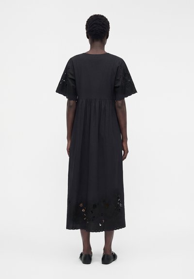 Femme vue de dos, portant une robe midi noire avec des manches courtes et des détails découpés floraux sur les manches et l'ourlet.