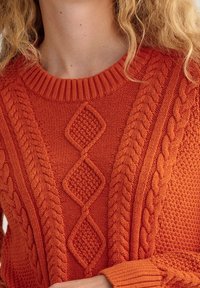 Pull tricoté orange avec un col rond, présentant des motifs texturés en diamant et en torsades sur le devant ainsi que des poignets côtelés.