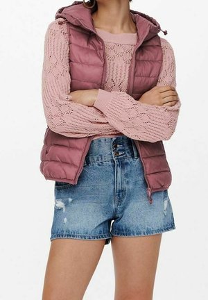 Vrouw draagt een mauve gebreide trui, bijpassend gewatteerd vest met capuchon en versleten blauwe denim shorts, met gekruiste armen over haar borst.