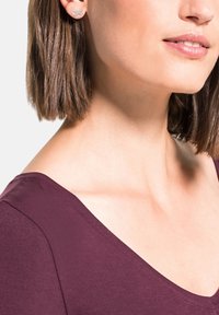 Donna con i capelli corti castani che indossa un top bordeaux e orecchini a stella di Argento a forma di infinito, sorridente.