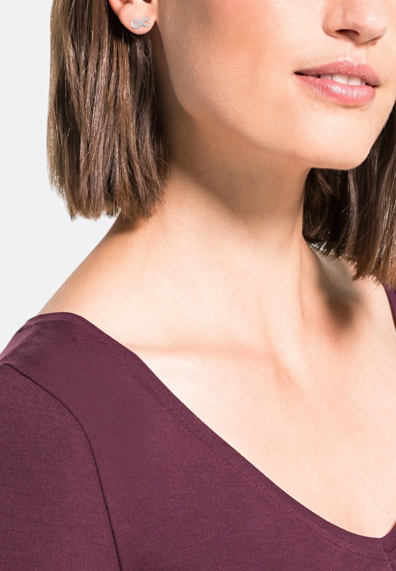 Donna con i capelli corti castani che indossa un top bordeaux e orecchini a stella di Argento a forma di infinito, sorridente.
