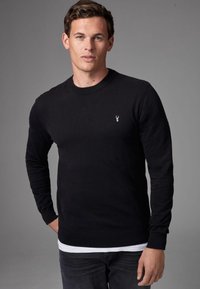 Pulover negru cu mânecă lungă și gât rotund, fabricat dintr-un material cu o textură fină; are un mic emblemă pe pieptul stâng.