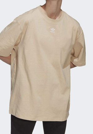 Person wearing oversized beige Adidas t-shirt with small white logo on chest, paired with black pants, standing with hands behind back.