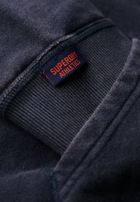 Ciemnoniebieski materiał bluzy o teksturowanym wykończeniu; zawiera czerwoną i granatową naszywkę "Superdry Athletics" wszytą w szew.