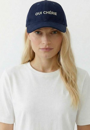 Frau mit langen blonden Haaren, die eine marineblaue Kappe mit der Aufschrift „OUI CHÉRIE“ und ein schlichtes weißes T-Shirt mit einem kleinen Herz-Emblem trägt.