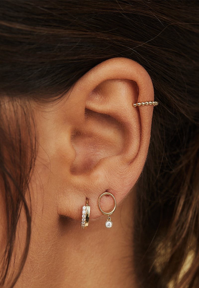 Gros plan d'une oreille avec trois boucles d'oreilles en or : une manchette perlée sur le haut de l'oreille, un petit anneau avec des pierres, et un anneau en or avec une pierre précieuse pendante.