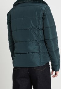 Teal pufferjacka med quiltad design, som har en fuskpälskrage, slät textur och en figursydd silhuett. Bakifrån syns längden som når till höfterna.
