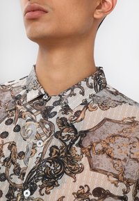 Camicia con colletto in tessuto leggero, caratterizzata da intricati motivi barocchi in tonalità marrone, crema e oro, con chiusura a bottoni e maniche corte.