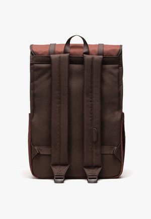 Ruskea kangaskassi, jossa on kääntöpäällys, varustettuna kahdella säädettävällä olkahihnalla ja ylhäällä olevalla kantokahvalla. Minimalistinen esteetti.