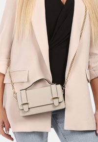 Beige handtas met gouden hardware, gestructureerd ontwerp, dubbele handvatten en een sluiting met haak. Gezet tegen een lichte blazer en een zwarte top als achtergrond.
