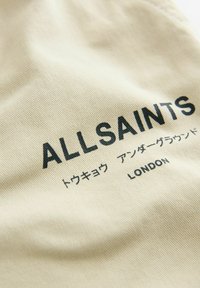 Beige stof med tekstureret overflade, der har "ALLSAINTS" logoet i fede sorte bogstaver, ledsaget af tekst på japansk og "LONDON" nedenunder.