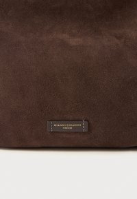 Braune Wildledertasche mit einem Lederlabel in Goldschrift, das eine glatte Textur und ein minimalistisches Design präsentiert.