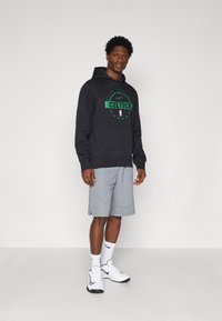 Čierna mikina s kapucňou a zeleným logom "Celtics", šedé športové kraťasy, biele tenisky a ponožky s čiernymi akcentmi. Jednoduchý, neformálny športový outfit.