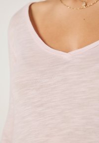 Svjetloružičasti top s V-izrezom izrađen od mekanog, teksturiranog materijala, s opuštenim krojem i suptilnim prugastim uzorkom. Bez vidljivih logotipa ili naglasaka.