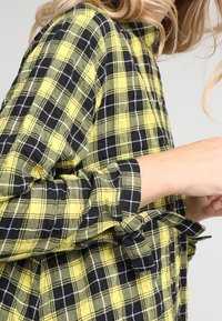 Camicia a quadri gialla e nera con polsino abbottonato, dettaglio cravatta in vita e un tessuto morbido e strutturato. Il motivo presenta controlli a contrasto.