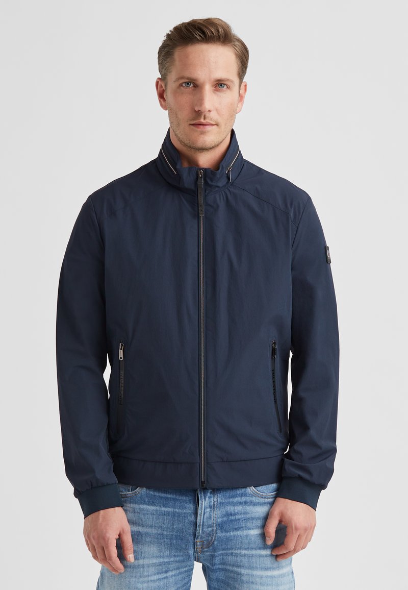 Marineblaue Jacke mit hohem Kragen, leichtem Stoff, seitlichen Taschen mit Reißverschlüssen und elastischen Bündchen. Glatte Textur, minimalistisches Design.