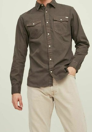 Homme portant une chemise marron à manches longues boutonnées avec poches poitrine et un pantalon beige clair, une main dans la poche, cadré au niveau du cou et des chevilles.