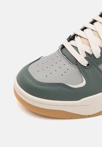 Baskets en cuir vert et gris avec une boîte à orteils perforée, des lacets beige et une semelle en caoutchouc gomme ; présente un design incurvé et des détails de couture.