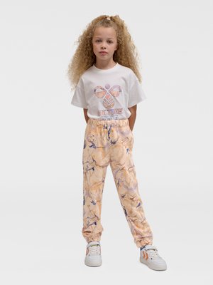 Junges Mädchen mit lockigem Haar, das ein weißes T-Shirt mit Grafik, pfirsichfarbene Hosen mit Marmormuster und weiße Sneaker mit orangefarbenen Akzenten trägt.