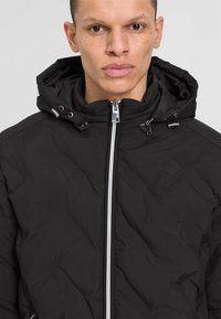 Schwarze Pufferjacke mit Reißverschluss vorne, verstellbarem Kapuze und strukturiertem Stoff. Mit silbernen Reißverschlüssen und Stoppern für einen sicheren Sitz.