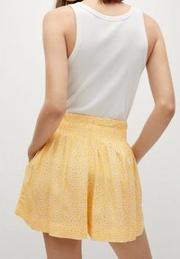 Shorts jaunes avec une taille élastique, ornés d'un motif floral et d'une texture de tissu léger. Portés avec un débardeur blanc.