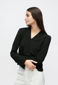 Melnas, garām piedurknēm top ar V veida izgriezumu un gludu tekstūru, kopā ar gaišas krāsas biksēm. Modelis pozē ar sakrustotām rokām.