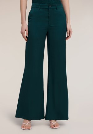 Donna che indossa pantaloni verde scuro a vita alta e gamba larga con bottoni frontali e cerniera, abbinati a sandali rosa chiaro con cinturini.