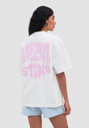 Femme aux longs cheveux foncés porte un t-shirt blanc oversize avec un grand texte rose « WESTBAY STUDIO » et un smiley dans le dos, short bleu clair.
