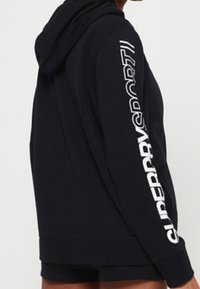 Sudadera negra con capucha y puños y dobladillo acanalados. Presenta un largo logo blanco "SUPERDRYSPORT" a lo largo de una manga. Tejido suave y texturizado.