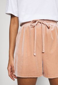 Primer plano de una persona vistiendo pantalones cortos de terciopelo rosa suave con cordón y camiseta blanca, mostrando un atuendo casual y relajado.