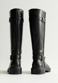 Botas de cuero negras altas con tacones gruesos y hebillas laterales, mostradas desde atrás sobre una superficie blanca y un fondo gris claro.