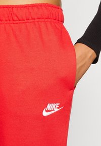 Červené bavlněné tepláky s bílým logem Nike na levém stehně. Mají elastický pas a boční kapsu. Hladká textura.