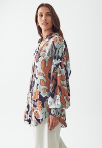 Blusa colorida con estampados florales en verde, naranja y blanco; mangas largas, cierre frontal de botones, dobladillo curvado y tejido ligero.