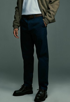 Marineblaue Hose in taillierter Passform, kombiniert mit einem braunen Gürtel und schwarzen Schuhen. Die strukturierte graue Jacke hat einen Reißverschluss und gerippte Bündchen.