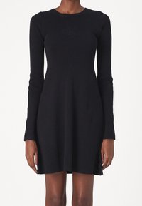 Femme portant une robe tricotée noire ajustée à manches longues avec un logo discret en relief sur la poitrine, debout devant un fond uni.