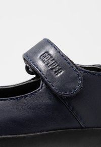 Zapato de cuero azul oscuro con punta redonda, una correa de velcro y detalles de costura en contraste. Textura suave y diseño minimalista.